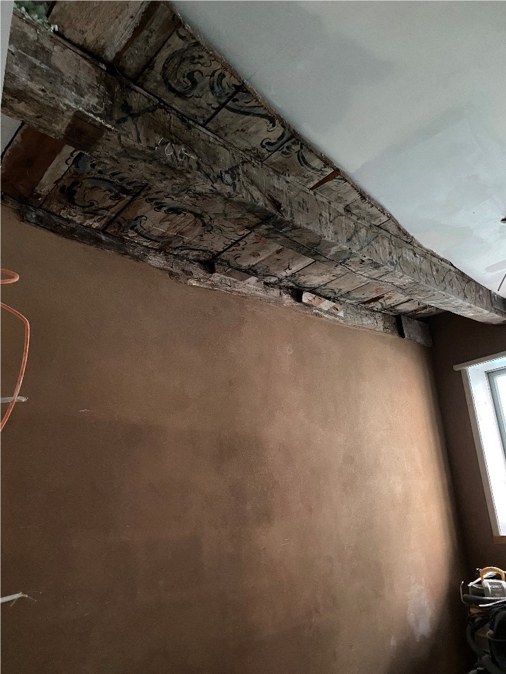 Mit Lehm überputzte Wand unter der bemalten Holzbalkendecke